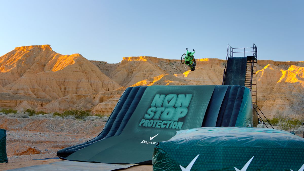 An individual using a green wheelchair attempts a flip above Degree branded crash pads.