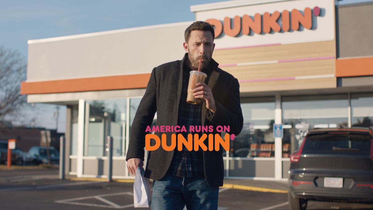 Actor Ben Affleck, wearing a blue plaid shirt and dark coat, stands in front of a Dunkin' sipping a coffee.